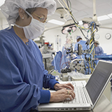 Nurse in full scrubs typing on a computer as doctors perform surgery in the background